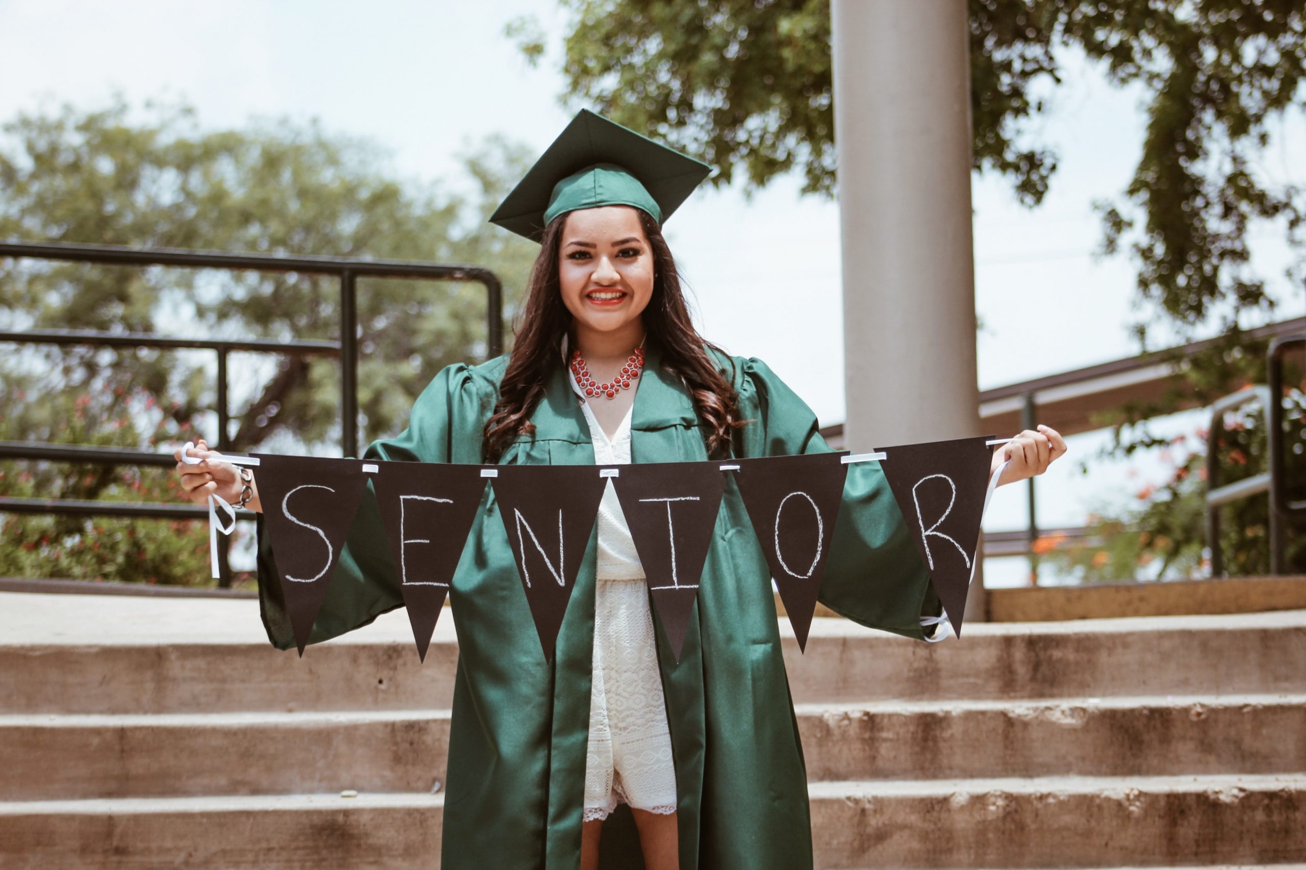 Image of teen in a green graduation cap and gown, holding a banner with "Seniors" on it. Photo by Juan Ramos on Unsplash
