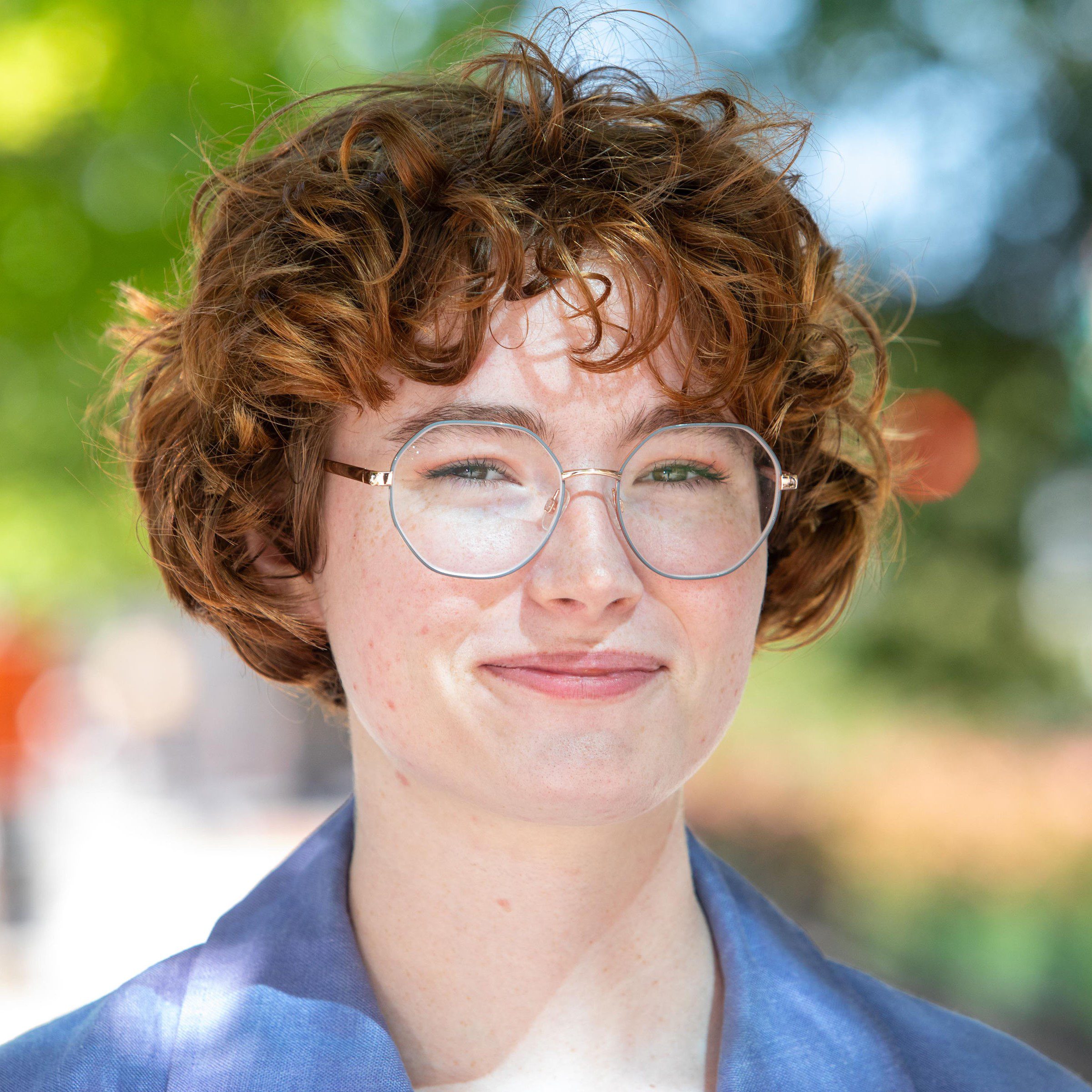 Photo of Bex Heimbrock from the shoulders up, facing the camera wearing a blue collared shir at round rimmed glasses.