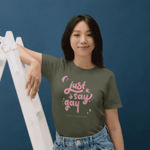 woman with short dark hair wearing an olive green colored t-shirt with pink script words that say "Just Say Gay"