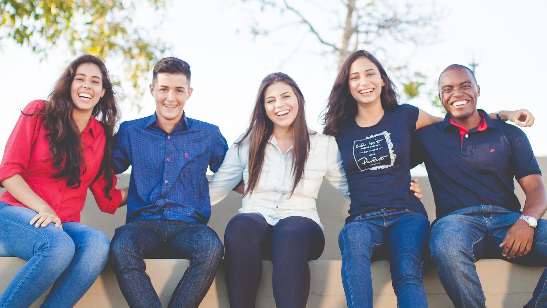 A group of young people sit on bench smiling sitting arm in arm