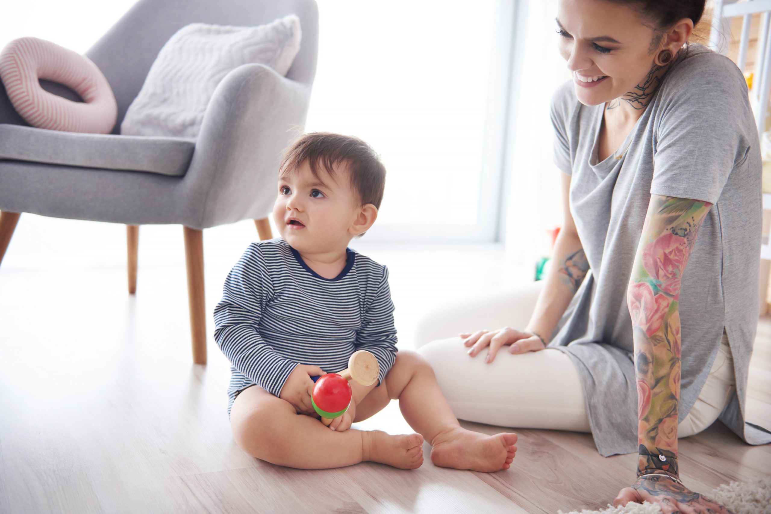 Photograph of a young parent sitting on the floor next to their baby, who is holding a toy
