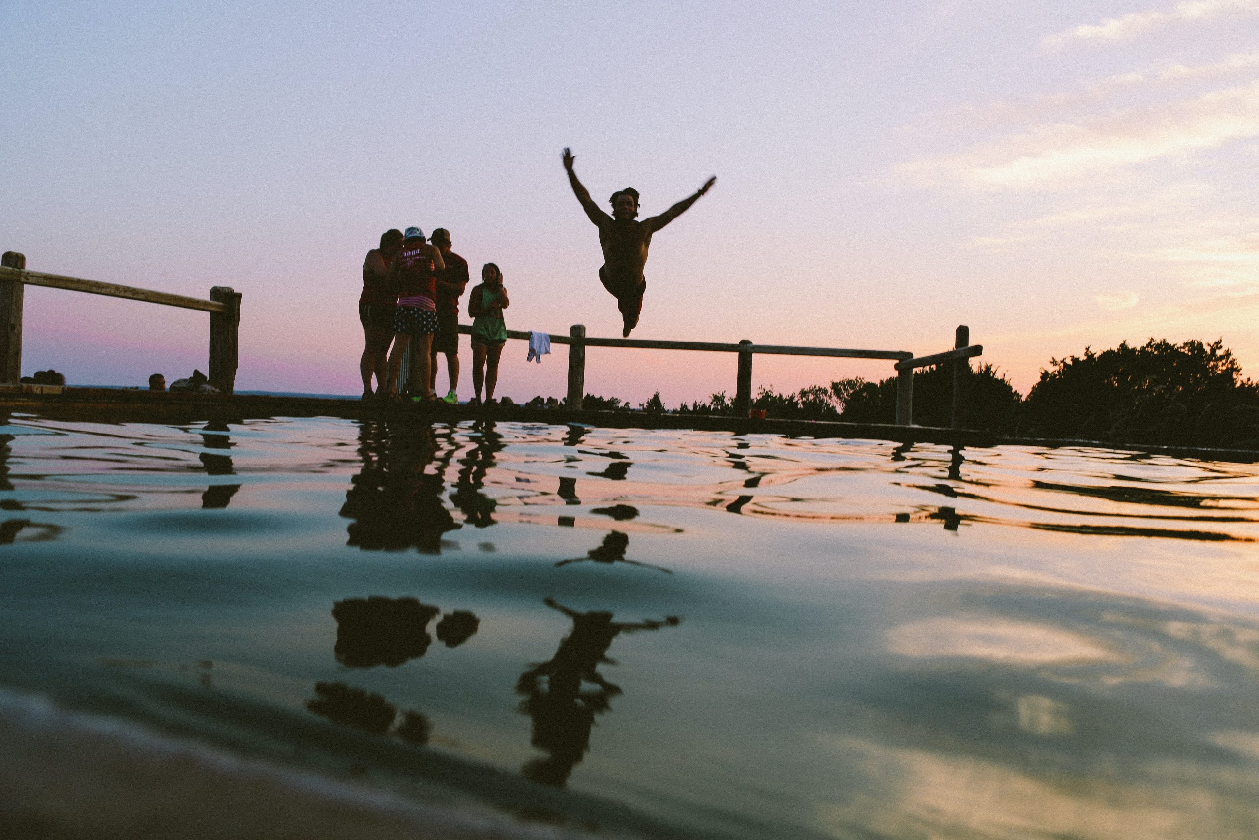 youth standing poolside at sunset with one person jumping in the pool
