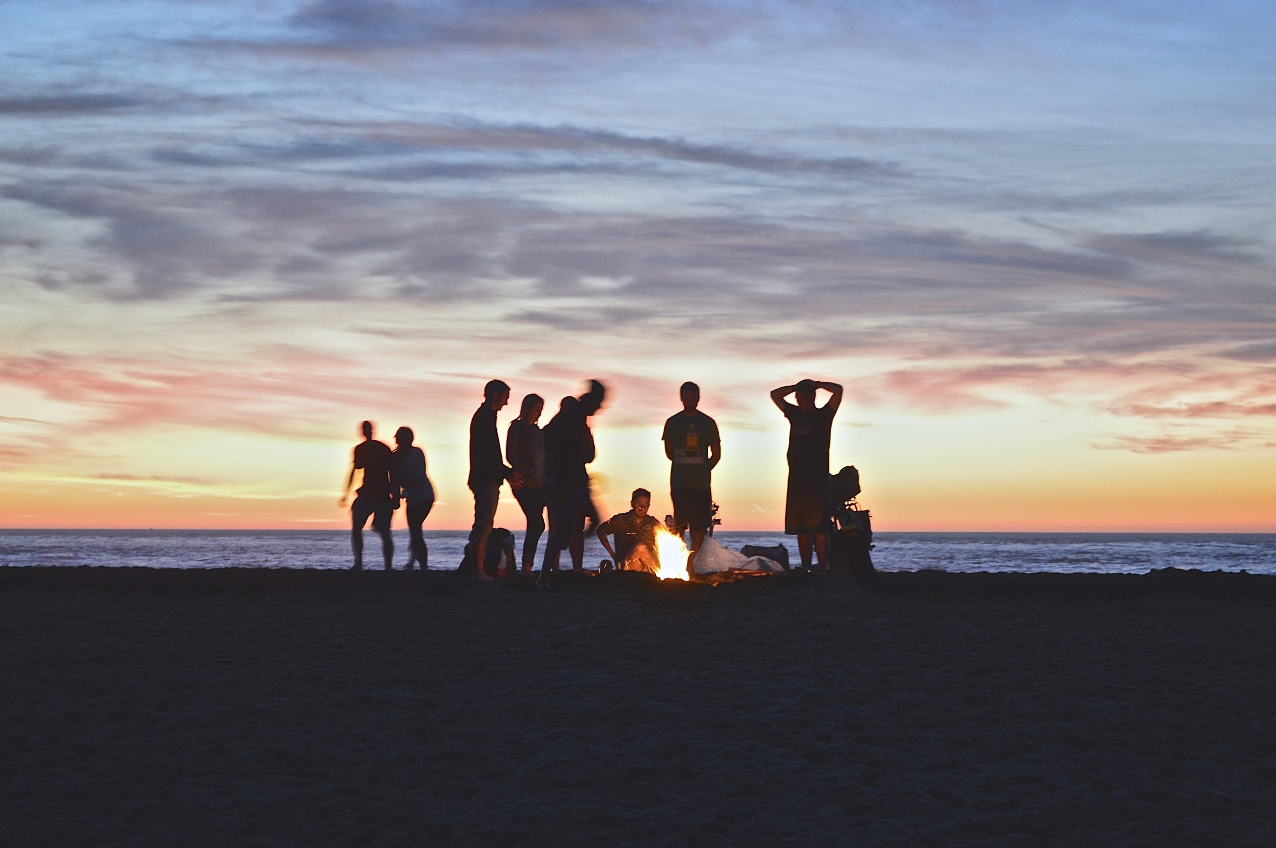 group of young people on the beach gathered around a bonfire at dusk